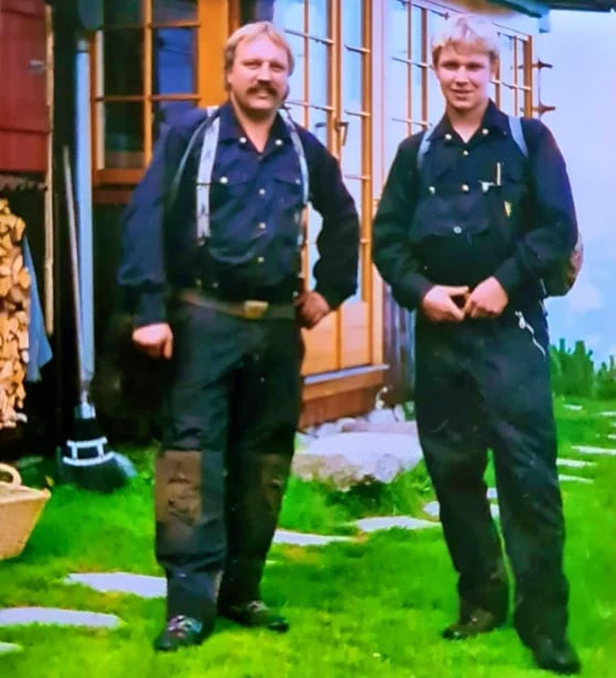 Zwei Männer in dunkler Arbeitsuniform und Overalls stehen auf einer Wiese vor einem Holzhaus mit orangefarbenem Besatz