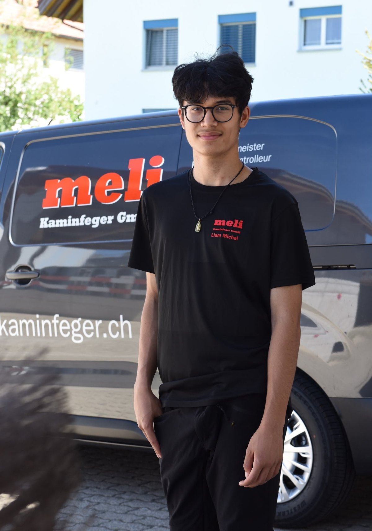 Junger Mann mit Brille in schwarzem Meli-Firmenshirt vor blauem Arbeitsfahrzeug